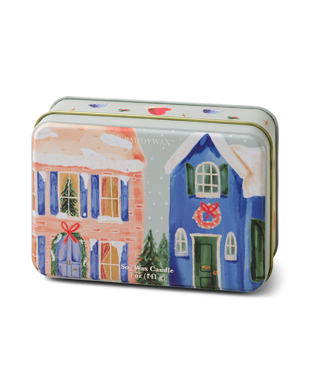 A rectangular tin candle with a painted winter scene featuring two houses. The house on the left is peach-colored with white windows and a red ribbon decoration, while the house on the right is blue with green windows and a wreath on the door. Snow is falling and accumulating on the roofs. The lid has an ornate gold-colored rim. The label on the front reads “Soy Wax Candle 14 oz.