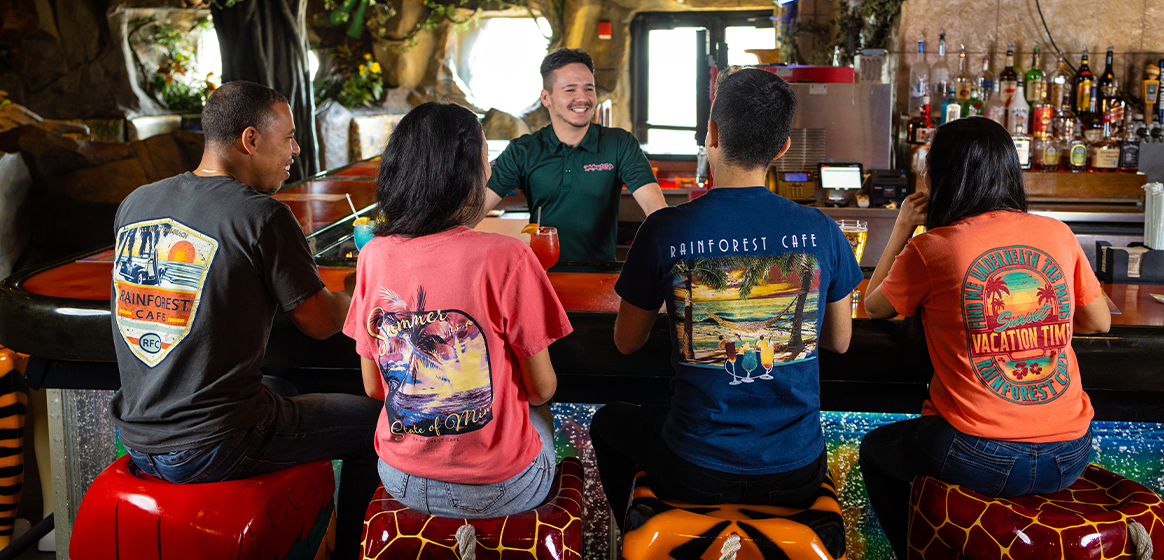 The image shows four individuals seated at a bar with their backs to the camera, wearing T-shirts with ‘Rainforest Cafe’ logos. Two shirts display vibrant animal graphics and the phrase ‘Vacation Time’. The bar is adorned with a colorful, mosaic-like surface and stocked with various bottles.
