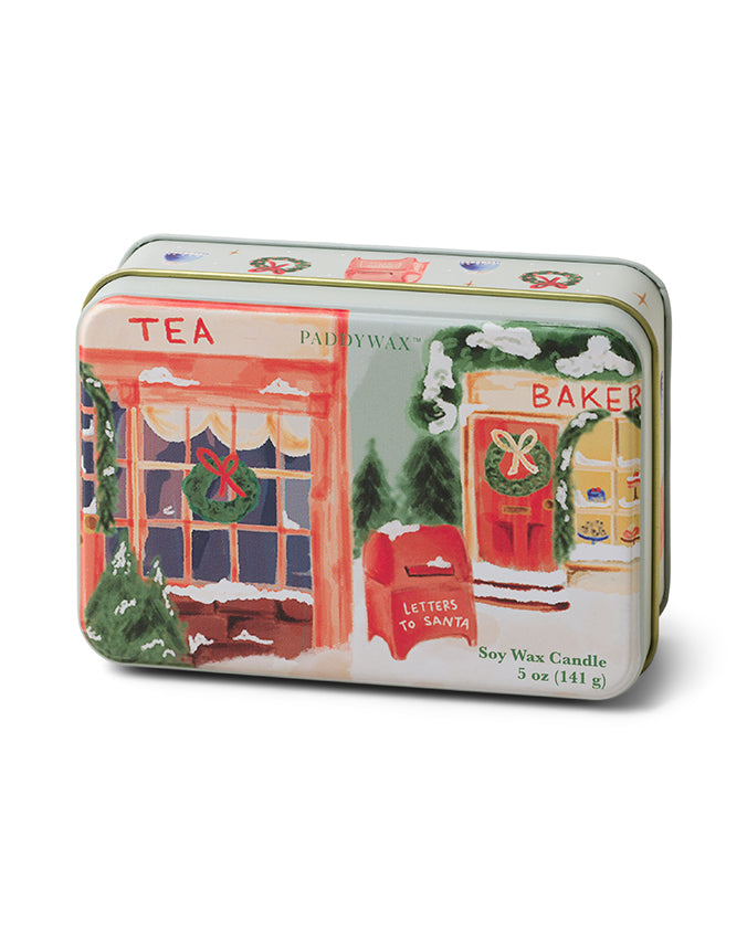 A rectangular tin soy wax candle with a festive design depicting a colorful storefront illustration. The left side of the tin shows a red and white striped awning above a window labeled “TEA,” adorned with a green wreath and red bow. The right side features an orange awning over a window labeled “BAKER,” also decorated with a wreath, next to which stands a red mailbox labeled “LETTERS TO SANTA.” Snow accents are visible on the awnings and mailbox, suggesting a winter holiday theme. The label on the tin stat