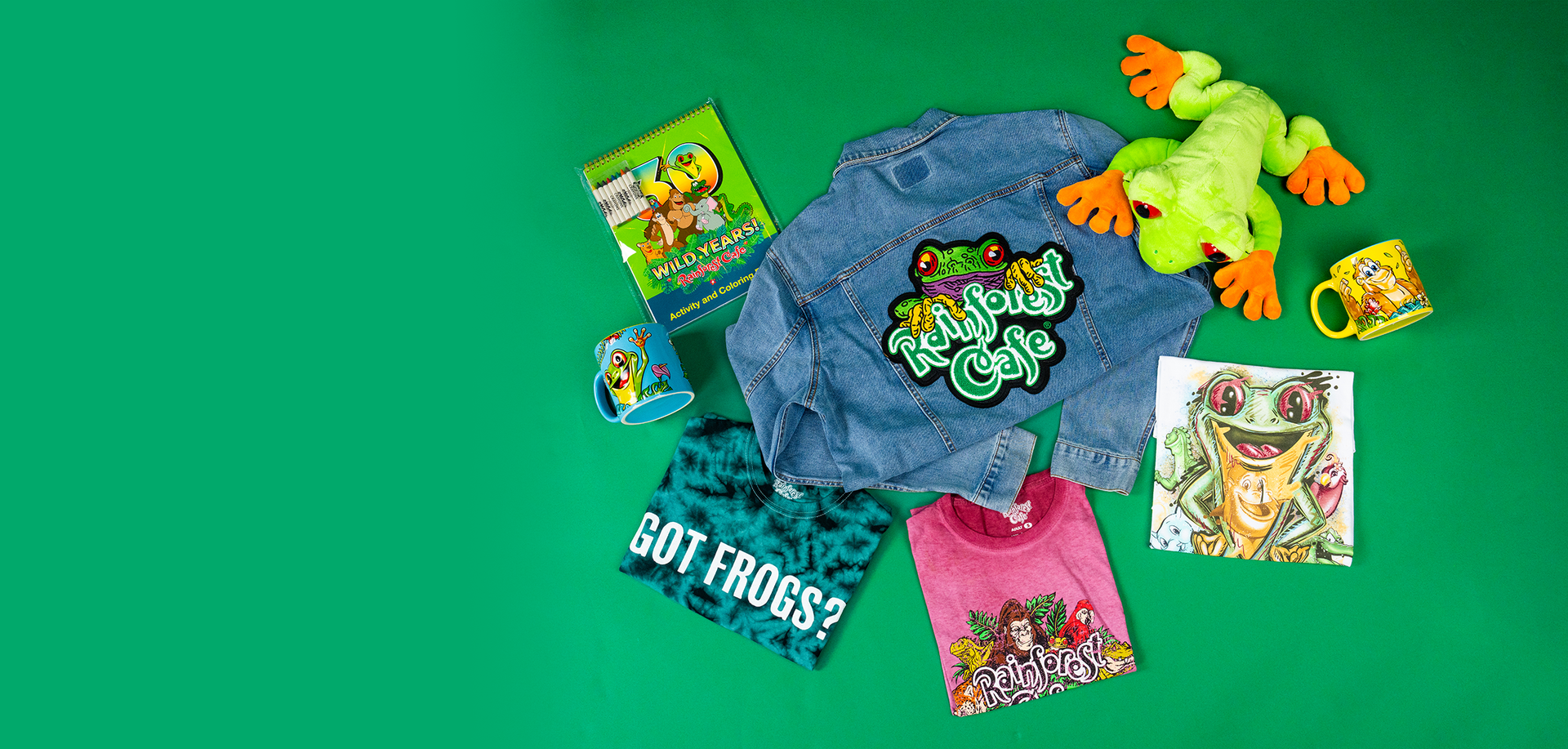 A collection of frog-themed items laid out on a green background. The items include a denim jacket with a large ‘Rainforest Cafe’ patch on the back, a plush green and yellow frog toy, two mugs with frog illustrations (one depicting a frog and the other showing a smiling oranguntang), a green tie-dye T-shirt with the text ‘GOT FROGS?’ in white letters, and a pink T-shirt featuring an illustration of frogs with the text ‘Rainforest Café.’ In the top left corner, there’s a small book or notebook with an image 