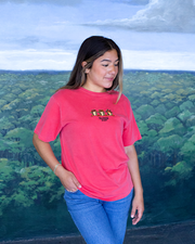 A person stands in front of a painted landscape, facing away from the camera. They are wearing a red t-shirt with yellow text and graphics, paired with blue jeans. The background painting depicts a green forest canopy under a light blue sky with scattered clouds.