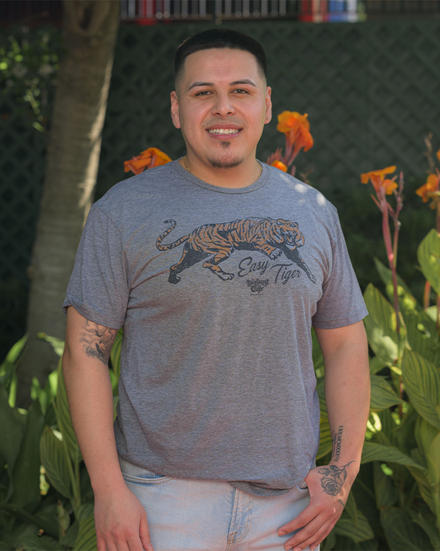 The image depicts a person standing in front of green foliage and orange flowers, wearing a grey t-shirt with a graphic of a tiger and the phrase “Easy Tiger” printed on it.