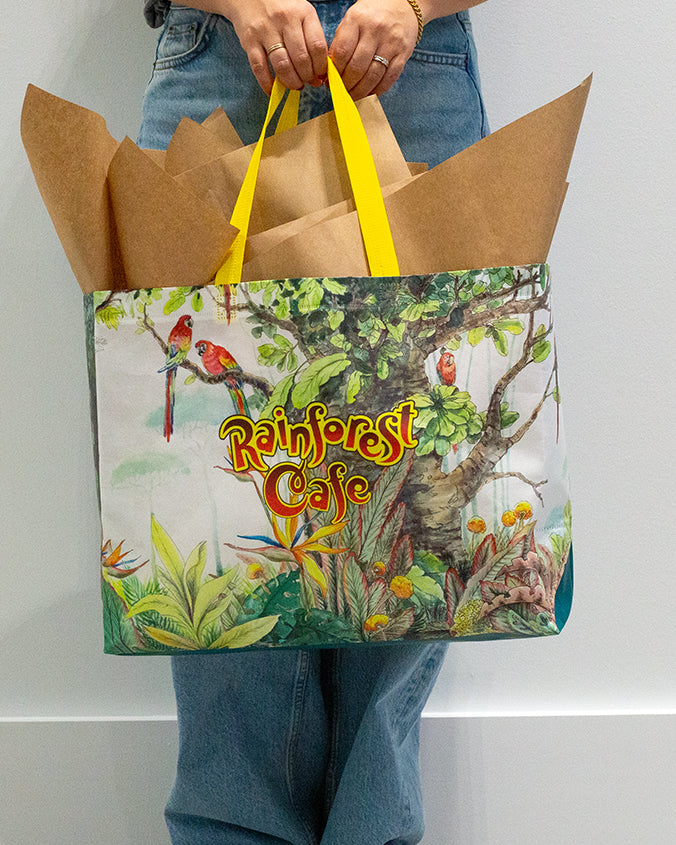 A lower-body view of a model in blue jeans holding a large, rainforest design featuring parrots on a tree, yellow handles, and the words "RAINFOREST CAFE" written in the middle. The background is plain white.