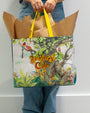 A lower-body view of a model in blue jeans holding a large, rainforest design featuring parrots on a tree, yellow handles, and the words "RAINFOREST CAFE" written in the middle. The background is plain white.