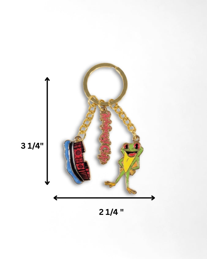 A gold-tone metal keychain with three dangling charms: a blue and red shoe charm, a pink script charm, and a green frog charm, shown on a white background with measurement arrows and labels indicating 3 1/4 inches tall by 2 1/4 inches wide.