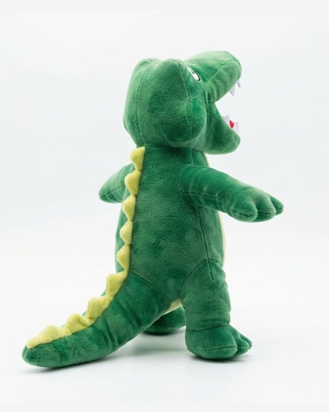 A green plush alligator toy standing on a white studio background, with yellow spikes along its back and tail, white teeth, and a small red tongue.