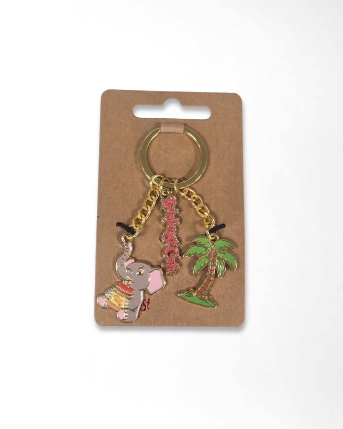 A gold-tone keychain with three charms displayed on a brown cardboard backing. The charms include a silver elephant with pink and yellow details, a pink rhinestone-encrusted word charm, and a green palm tree.