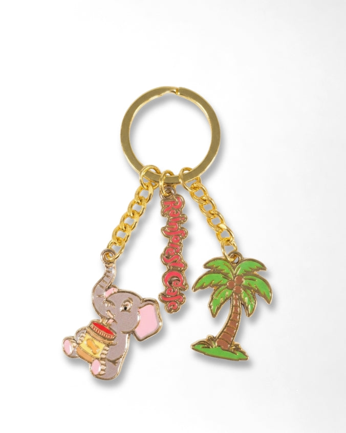 A gold-tone metal keychain featuring three charms: a pink elephant holding a yellow drum, a vertical red Chinese character charm, and a green palm tree charm, all attached to a gold split ring on a white background.