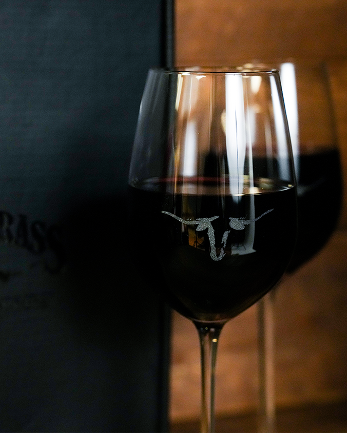 Two wine glasses filled with red wine beside a black box. One glass is in front of the other showcasing a longhorn print design in the middle.