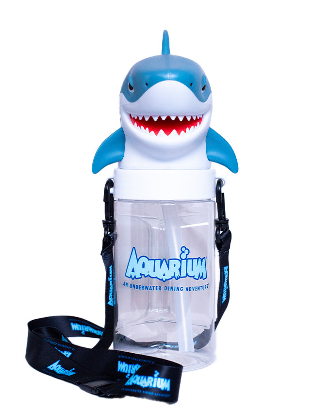 A novelty clear plastic backpack with a blue and white shark design protruding from the top. The shark has a wide, cartoonish grin with pointed teeth. The backpack is printed with the text ‘Aquarium An Underwater Dining Adventure’ in blue lettering. It has black straps with the word ‘Aquarium’ repeated along their length in white text.