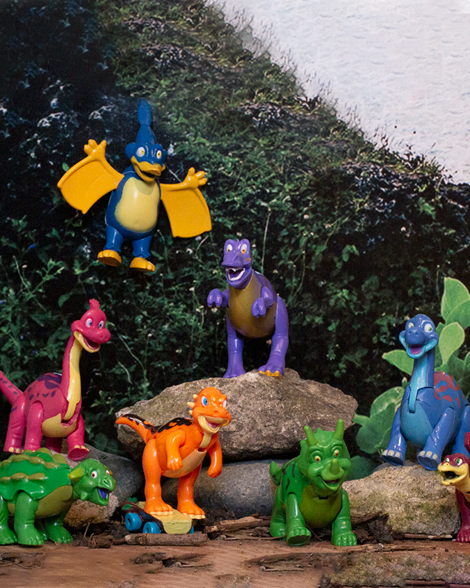 Eight colorful toy dinosaurs in front of a natural setting with rocks and bushes. The dinosaurs are of various species and colors.