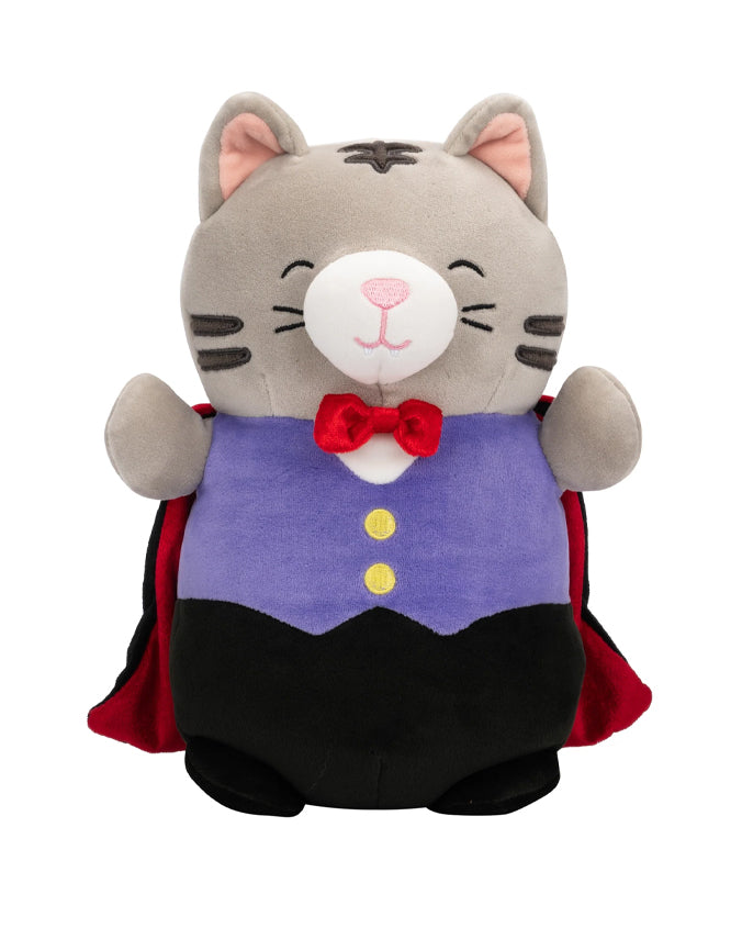 A plush toy resembling a cat standing upright. The toy has a gray and white face with whiskers, closed eyes, and a smiling expression. It wears a purple vest with two yellow buttons over a white shirt, complete with a red bow tie. The cat also has a black cape draped over its shoulders.