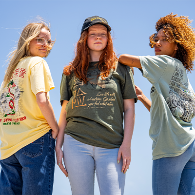 Three individuals standing side by side against a clear blue sky. The person on the left is wearing a yellow t-shirt with red text and graphics, the middle person is in a green t-shirt with gold text and graphics, and the person on the right is in a light blue t-shirt with black text and graphics. All are in denim pants.