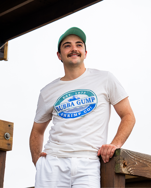 A young man stands on a pier against a white sky with his hand in his white bottoms pocket, wearing a white crewneck shirt with a circular blue logo reading "EST. 1975 BUBBA GUMP SHRIMP CO." with a ship inside. He is also wearing a teal baseball cap. 