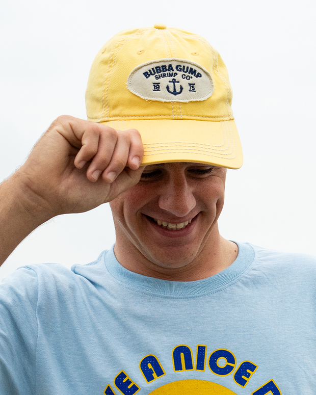 A headshot of a smiling model holding and wearing a yellow baseball cap with a frayed circular logo reading "BUBBA GUMP SHRIMP CO." with an anchor symbol and 19 75 under it. The person is wearing a blue shirt with bold text on it.