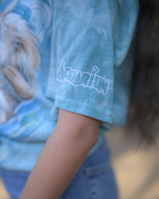 A close-up view of a person’s arm wearing a blue tie-dye shirt with the word ‘Aquarium’ in white stylized text on the sleeve. The swirling patterns of the tie-dye are visible, and the background is out of focus.