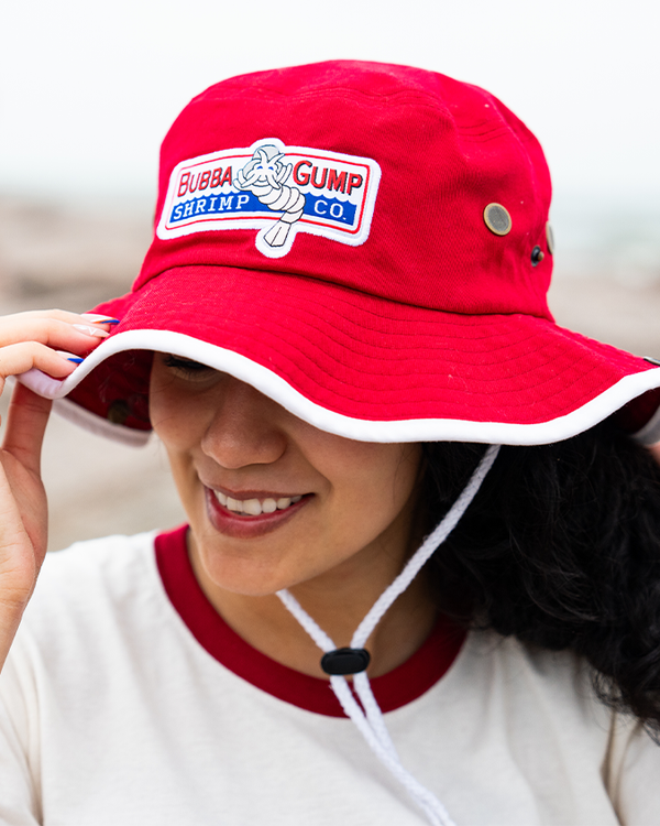 A headshot of a dark-haired model wearing a white and red collared crewneck shirt and holding onto and wearing a red bucket hat with white outlining and adjustable strings, as well as a rectangular "Bubba Gump Shrimp Co." logo, with a shrimp on it. The background is plain and blurred.