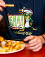 A close-up mid-level view of a model at a table eating French fries, wearing a navy-blue t-shirt with an illustration of a cartoon frog in a chef outfit holding a burger and fries, along with the text "CHA! CHA! burgers & fries Rainforest Cafe" in a wooden frame. "RAINFOREST CAFE" is written under it in stylized font.