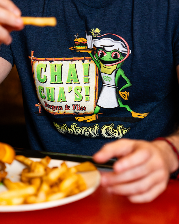A close-up mid-level view of a model at a table eating French fries, wearing a navy-blue t-shirt with an illustration of a cartoon frog in a chef outfit holding a burger and fries, along with the text "CHA! CHA! burgers & fries Rainforest Cafe" in a wooden frame. "RAINFOREST CAFE" is written under it in stylized font.