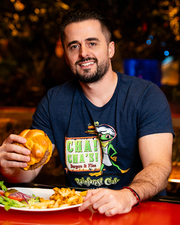 A model sitting at a table inside of a restaurant holding a burger in one hand, with a plate of food in front of them. The model is wearing a navy-blue t-shirt with an illustration of a cartoon frog in a chef outfit holding a burger and fries, along with the text "CHA! CHA! burgers & fries Rainforest Cafe" in a wooden frame. "RAINFOREST CAFE" is written under it in stylized font.
