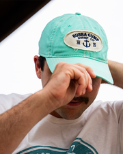 A headshot of a model wearing a white shirt, adjusting and wearing a teal baseball cap with a frayed circular logo reading "BUBBA GUMP SHRIMP CO." with an anchor symbol and 19 75 under it.