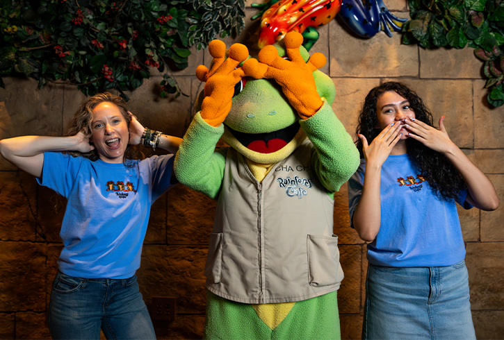 Two smiling individuals in blue shirts pose playfully with a cheerful green frog mascot at a themed restaurant backdrop.