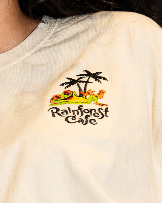 A close-up shoulder-level view of a model wearing a beige-colored T-shirt with a small graphic design on the left chest area featuring two black palm trees, a relaxing frog, and the text "Rainforest Cafe" written in black a stylized font.