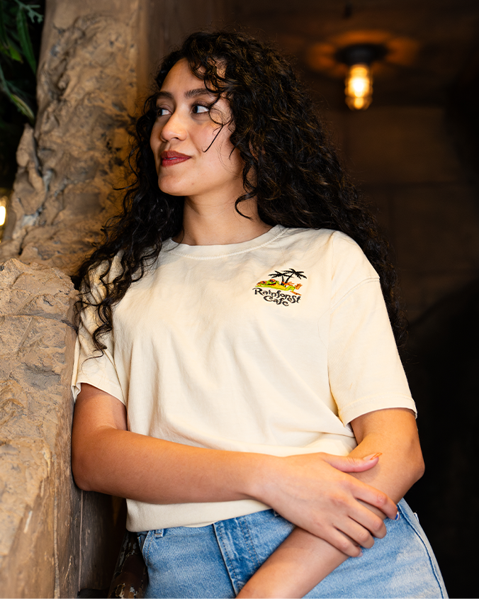 A waist-up view of a dark-haired model with one hand holding her forearm, wearing a beige-colored T-shirt with a small graphic design on the left chest area featuring two palm trees, a relaxing laying frog, and the text "Rainforest Cafe" written in a stylized font. The model is leaning against a rock surface in a dimly lit room.