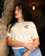 A waist-up view of a dark-haired model with one hand holding her forearm, wearing a beige-colored T-shirt with a small graphic design on the left chest area featuring two palm trees, a relaxing laying frog, and the text "Rainforest Cafe" written in a stylized font. The model is leaning against a rock surface in a dimly lit room.