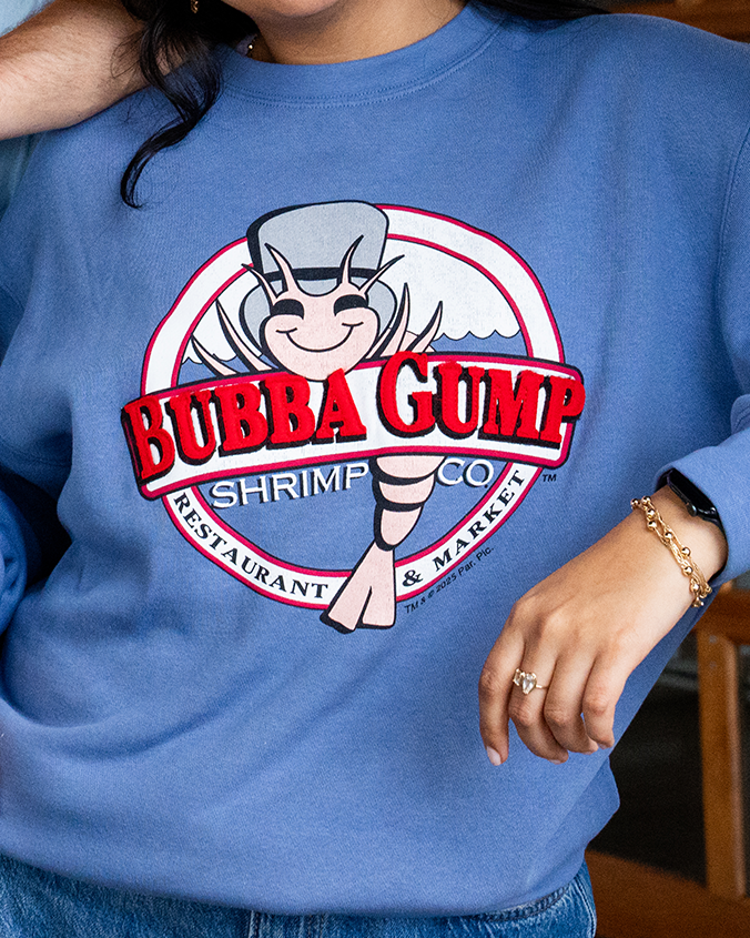 A chest-level view of a model wearing a blue crewneck sweatshirt featuring a circular Bubba Gump Shrimp Co. logo in the center, with blue jeans and gold jewelry.