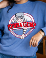 A chest-level view of a model wearing a blue crewneck sweatshirt featuring a circular Bubba Gump Shrimp Co. logo in the center, with blue jeans and gold jewelry.