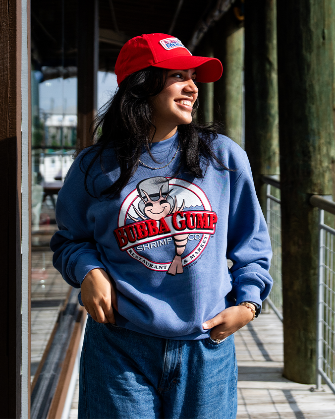 A waist-up view of a model walking on a wooden pier with her hands in her pockets, wearing a blue crewneck sweatshirt featuring a circular Bubba Gump Shrimp Co. logo, paired with denim jeans and a red cap with an embroidered logo in the center.