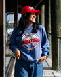 A waist-up view of a model walking on a wooden pier with her hands in her pockets, wearing a blue crewneck sweatshirt featuring a circular Bubba Gump Shrimp Co. logo, paired with denim jeans and a red cap with an embroidered logo in the center.