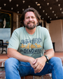 A brunette bearded model sitting on brick steps in front of a building, wearing blue jeans, a watch and a light green crewneck T-shirt with a design on the front featuring four dinosaur-shaped chicken nuggets and text that reads: "Never TOO OLD for DINO NUGS - T-REX ORLANDO". 