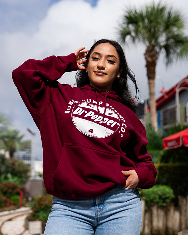 A model poses confidently in a maroon hoodie with a Dr. Pepper graphic, a front pocket and drawstrings. The hoodie has a circular logo on the chest area that reads "Bubba Gump Shrimp Co." at the top, "Dr Pepper" in the middle, and "10 2 4" in between. set against a backdrop of palm trees and urban scenery.
