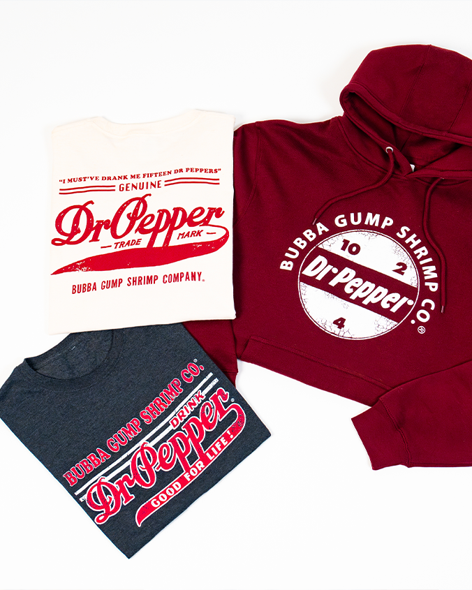Three articles of clothing with Dr Pepper and Bubba Gump Shrimp Co. branding. A white T-shirt reads 'I MUST'VE DRANK ME FIFTEEN DR PEPPERS' and 'GENUINE Dr Pepper TRADE MARK BUBBA GUMP SHRIMP COMPANY', next to a maroon hoodie featuring a circular logo and the numbers 10, 2, and 4. At the bottom is a dark gray T-shirt with a linear design reading "GOOD FOR LIFE!" at the bottom. The background is plain white.