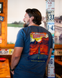 A backside view of a model inside of a rustic restaurant setting, wearing a blue crewneck T-shirt with a graphic on the back featuring an illustration of a long road in the canyons, with the text “World Famous GUMP HILL” at the top. Below the image are geographic coordinates. 