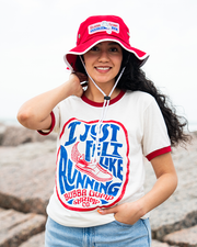A waist-up view of a dark-haired model in front of a rocky beach with her hand on her neck and one hand tucked into blue jeans, wearing a red bucket hat with white details and adjustable string, including a rectangular Bubba Gump Shrimp Logo. She is also wearing a white baseball t-shirt with red cuff lines, featuring a retro circular design with blue and red wording reading "I Just Felt Like Running" and "Bubba Gump Shrimp Co.", and a shoe with a feather in the middle. 