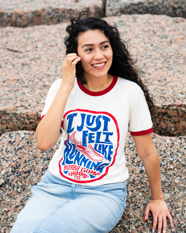 A knee-up view of a dark-haired model sitting on a marble rock with her hand touching her hair, wearing a jean skirt, and a white baseball t-shirt with red cuff lines, featuring a retro circular design with blue and red wording reading "I Just Felt Like Running" and "Bubba Gump Shrimp Co.", and a shoe with a feather in the middle.
