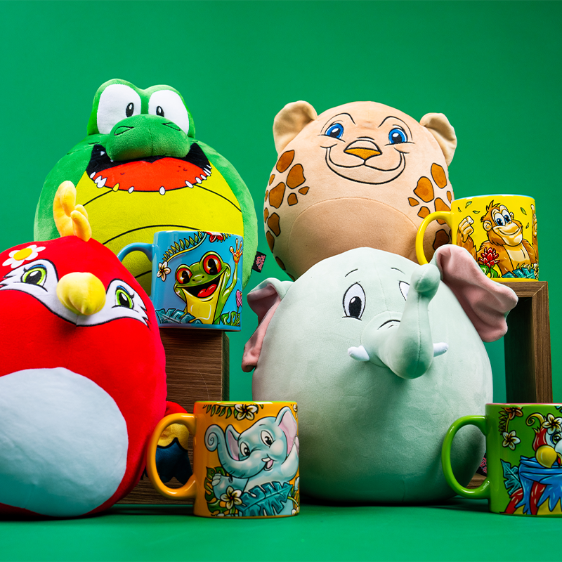 A collection of colorful plush toys and printed mugs on a green background. The plush toys include a red bird, a green frog with yellow details, a tan jaguar with brown ears, and a gray elephant with large ears. Each toy has cartoon-like features and expressions. There are also four mugs, each with an illustration matching the animals of the plush toys. The items are arranged in and around a wooden box, creating an engaging display of vibrant colors and patterns that draw attention to the playful nature of 