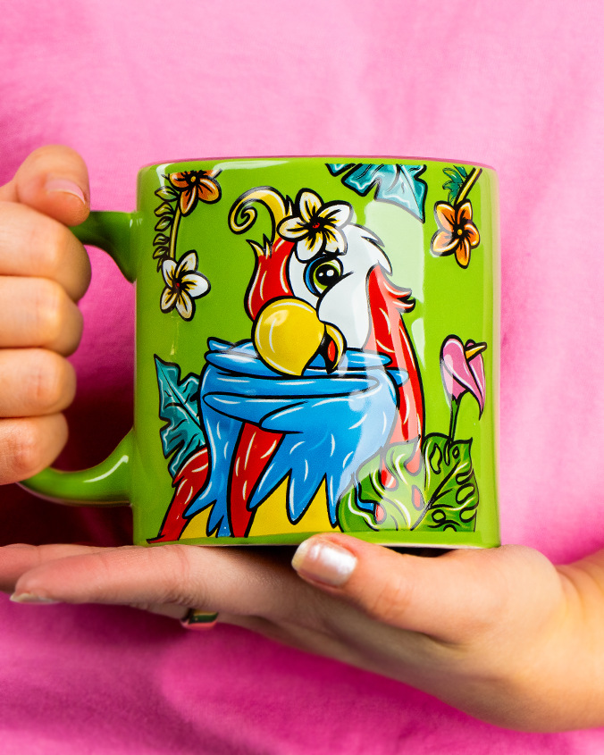 A close-up view of a model in a pink shirts' hands holding a colorful green mug with a posing parrot surrounded by tropical flowers and leaves.