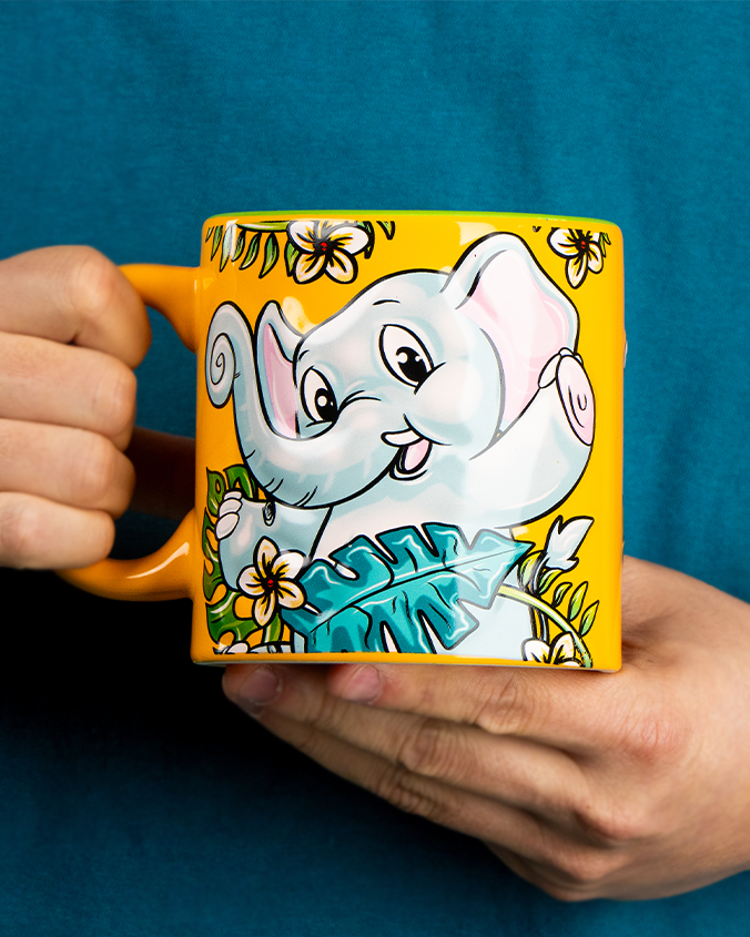 A close-up view of a model in a blue shirts' hands holding a colorful yellow mug with a posing elephant surrounded by tropical flowers and leaves. 