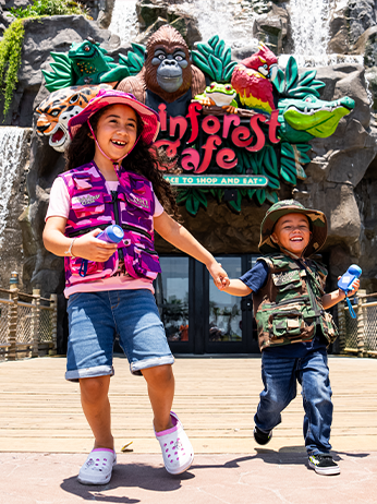 Two children are holding hands in front of the entrance to a themed cafe. The entrance is adorned with vibrant green foliage and features a large, red-eyed tree frog statue above the sign that reads ‘Rainforest Cafe.’ The child on the left wears a pink vest and white shoes, while the child on the right is dressed in camouflage clothing and blue shoes. They appear to be in motion, possibly walking or dancing.