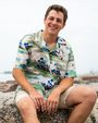 A brunette model sitting on a rock with his hands resting on his legs in front of a beach, wearing a colorful tropical shirt with tropical island and boats on it, and khaki shorts. 