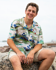 A brunette model sitting on a rock with his hands resting on his legs in front of a beach, wearing a colorful tropical shirt with tropical island and boats on it, and khaki shorts. 