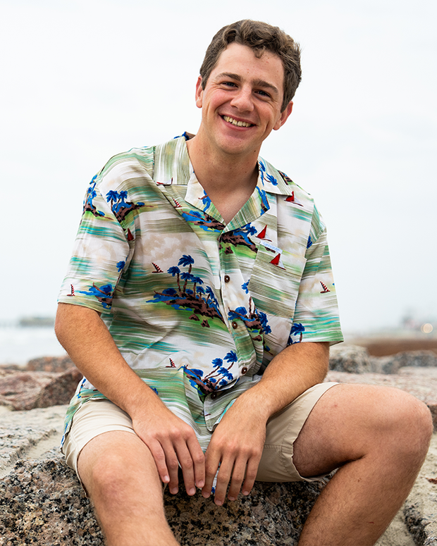 A brunette model sitting on a rock with his hands resting on his legs in front of a beach, wearing a colorful tropical shirt with tropical island and boats on it, and khaki shorts. 