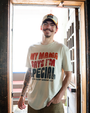 A waist-up view of a tattooed model standing in front of an open wooden door with his hand in his jean pocket, wearing a cream-colored shirt that reads "MAMA SAYS I'M SPECIAL, BUBBA GUMP SHRIMP CO." in the center. He is also brown and white baseball cap with the same wording. 