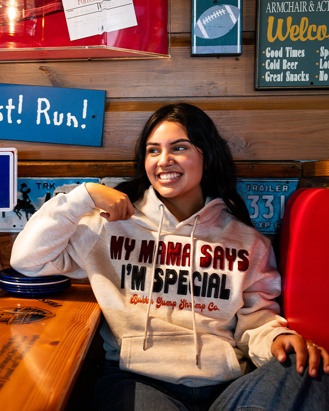 A dark-haired model sitting in a wooden booth in a rustic restaurant wearing blue jeans and a grey hoodie with text reading "MY MAMA SAYS I'M SPECIAL" in red and navy patched lettering, and "Bubba Gump Shrimp Co. in orange under it. 