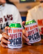 A zoomed in image of two models' at a wooden table, holding green cans inside of beige koozies with the text "MY MAMA SAYS I'M SPECIAL" and "BUBBA GUMP SHRIMP CO" printed on it in faded orange, red and black letters. 