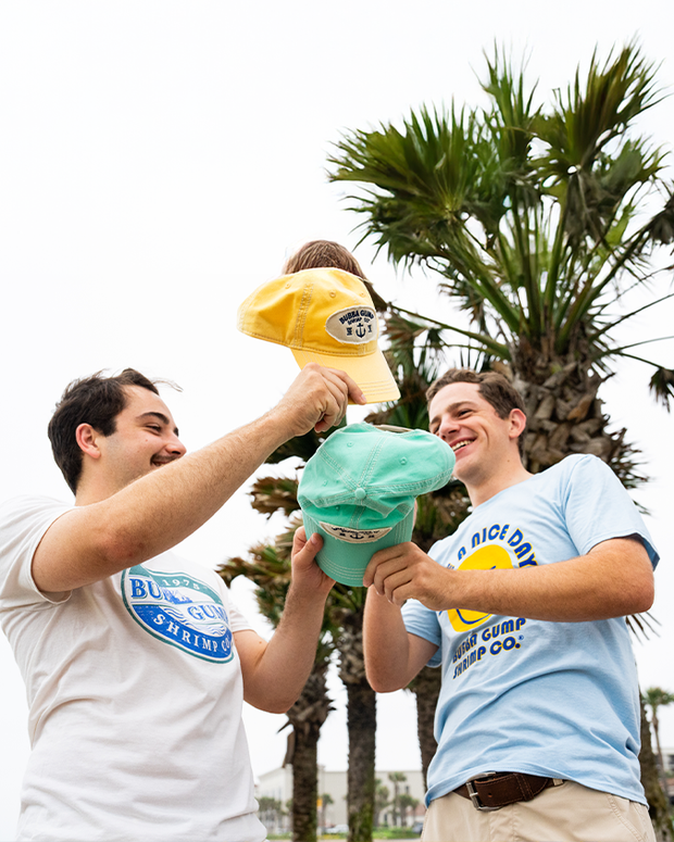 Two friends outside in front of palm trees holding teal and yellow hats with white circular patches with navy lettering and anchors on them. They are wearing colorful graphic shirts with circular logos.